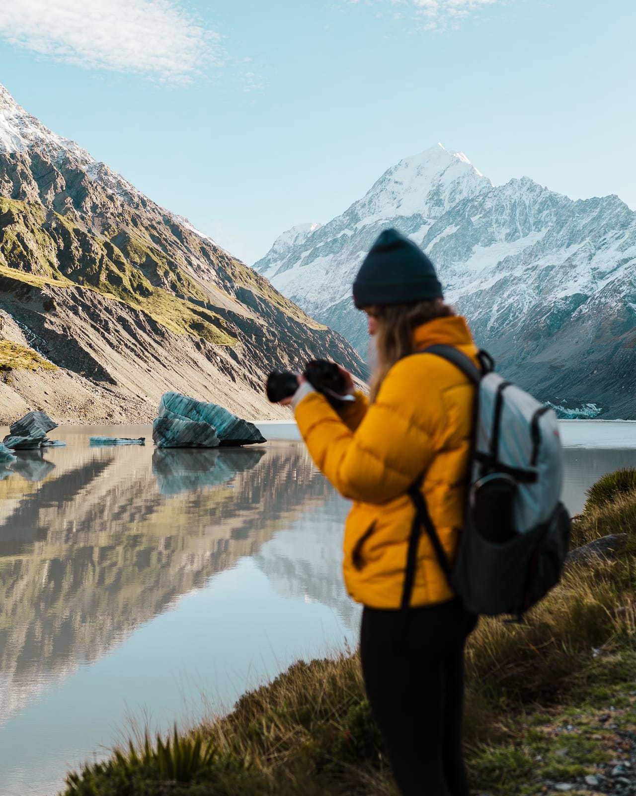 woman standing next to lake with ice berg and mountains in hooker valley, new zealand